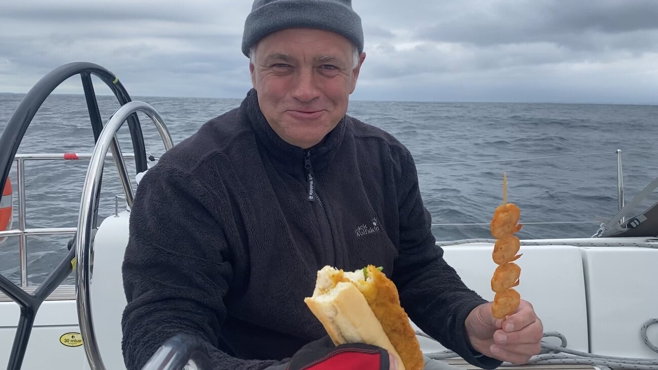 Ein Mann hält zwei Snacks in der Hand und lächelt in die Kamera während er vor dem Steuerrad sitzt